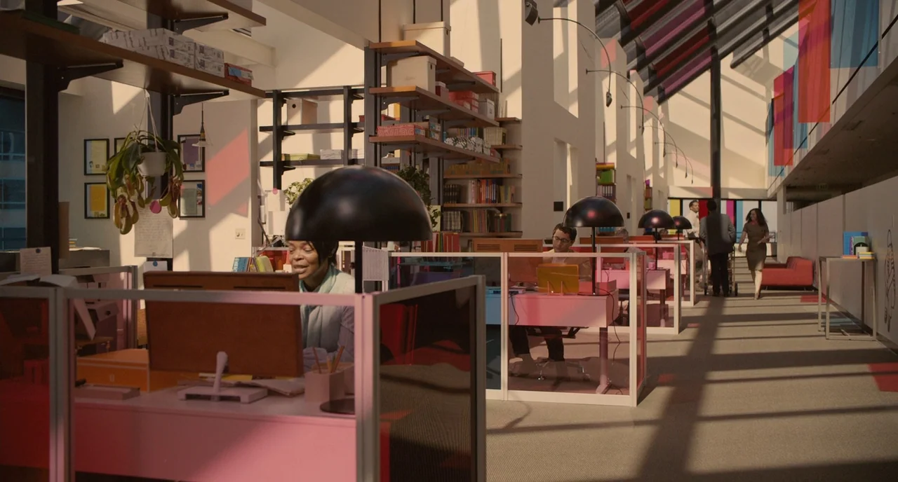 Her (2013) movie still: wide — A bright open plan workspace features rows of cubicles under a high, sloped ceiling. Multiple employees work at their…
