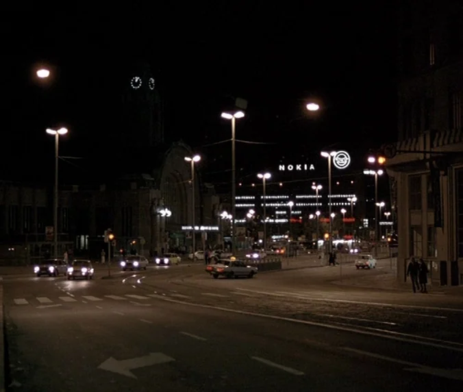 Crime & Punishment (1983) movie still: establishing — A city street intersection at night features a large clock building and signage. Cars with…