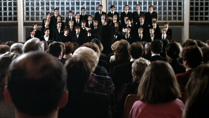 Benny’s Video (1992) movie still: wide — A large choir of boys dressed in formal suits stands on elevated risers. A conductor stands before them in…