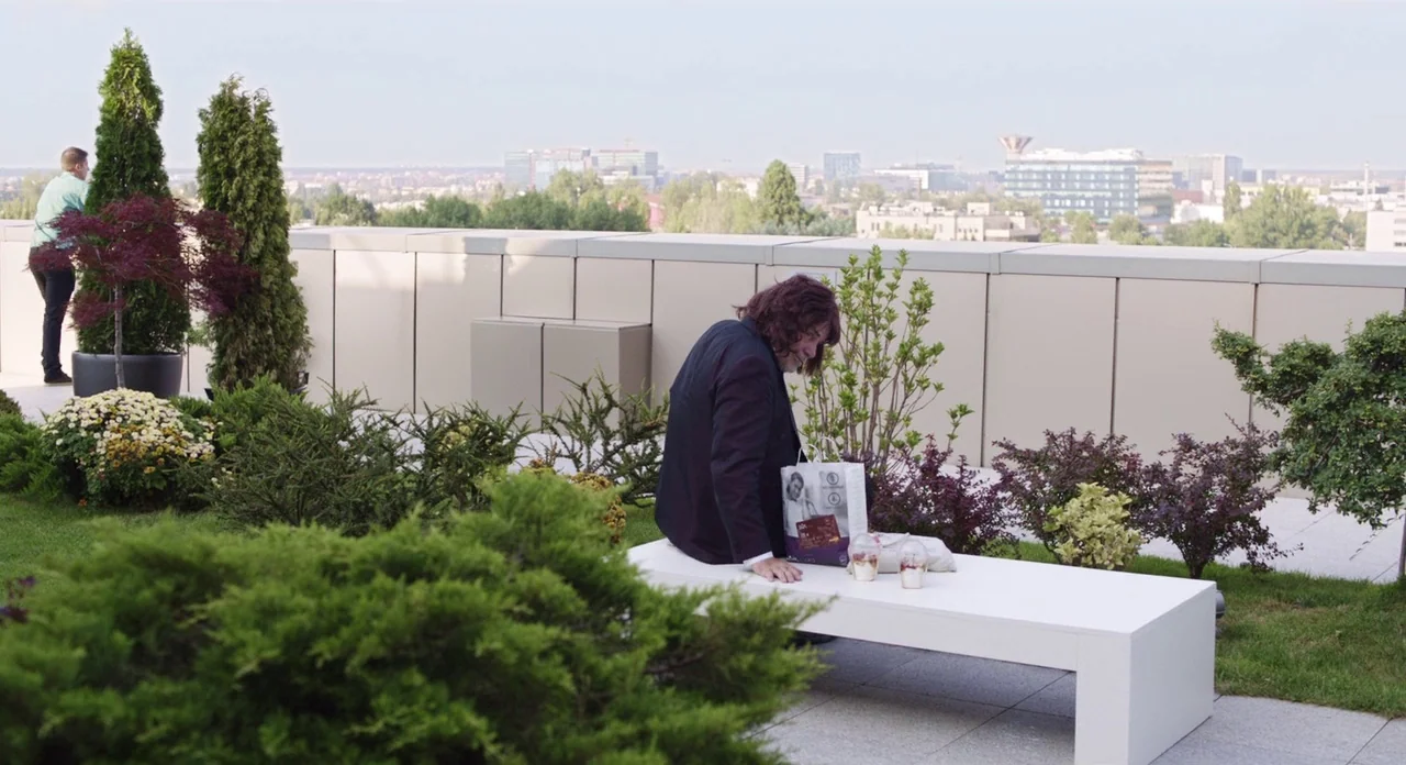 Toni Erdmann (2016) movie still: wide — A flat rooftop terrace features white benches, trimmed evergreen bushes, and a city view. A woman sits on a…