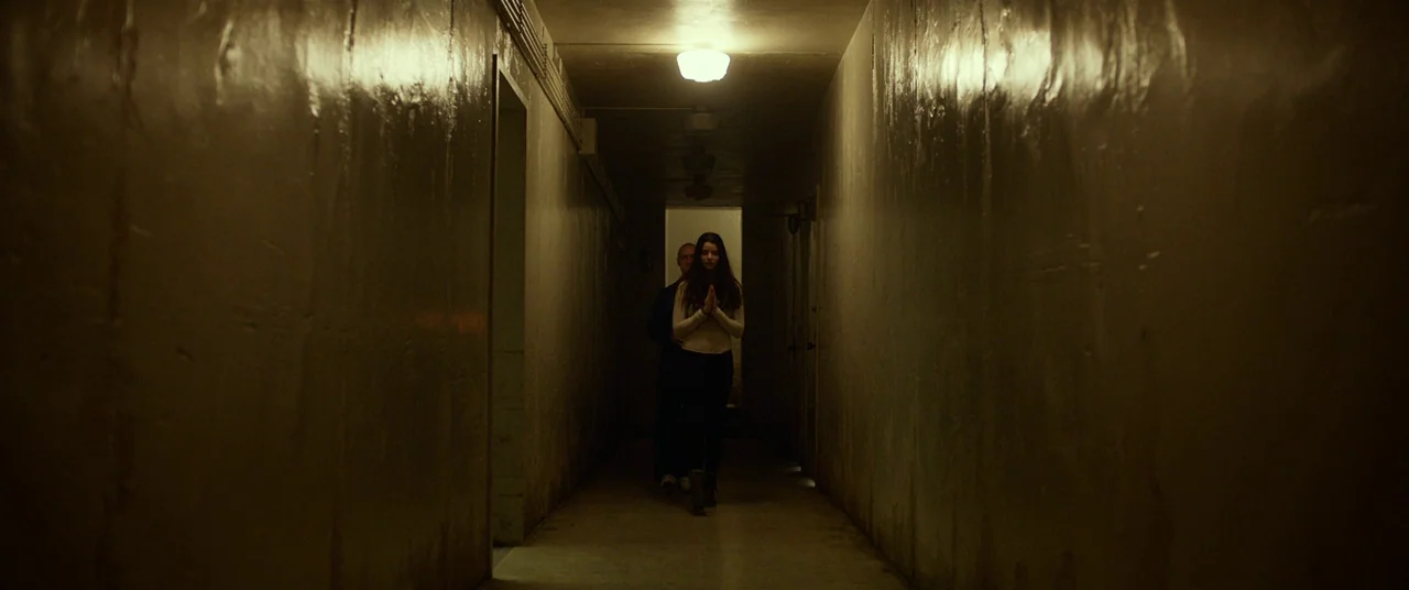 Split (2016) movie still: wide — A narrow, dimly lit industrial hallway features smooth, monolithic walls and a single overhead light. A young woman…