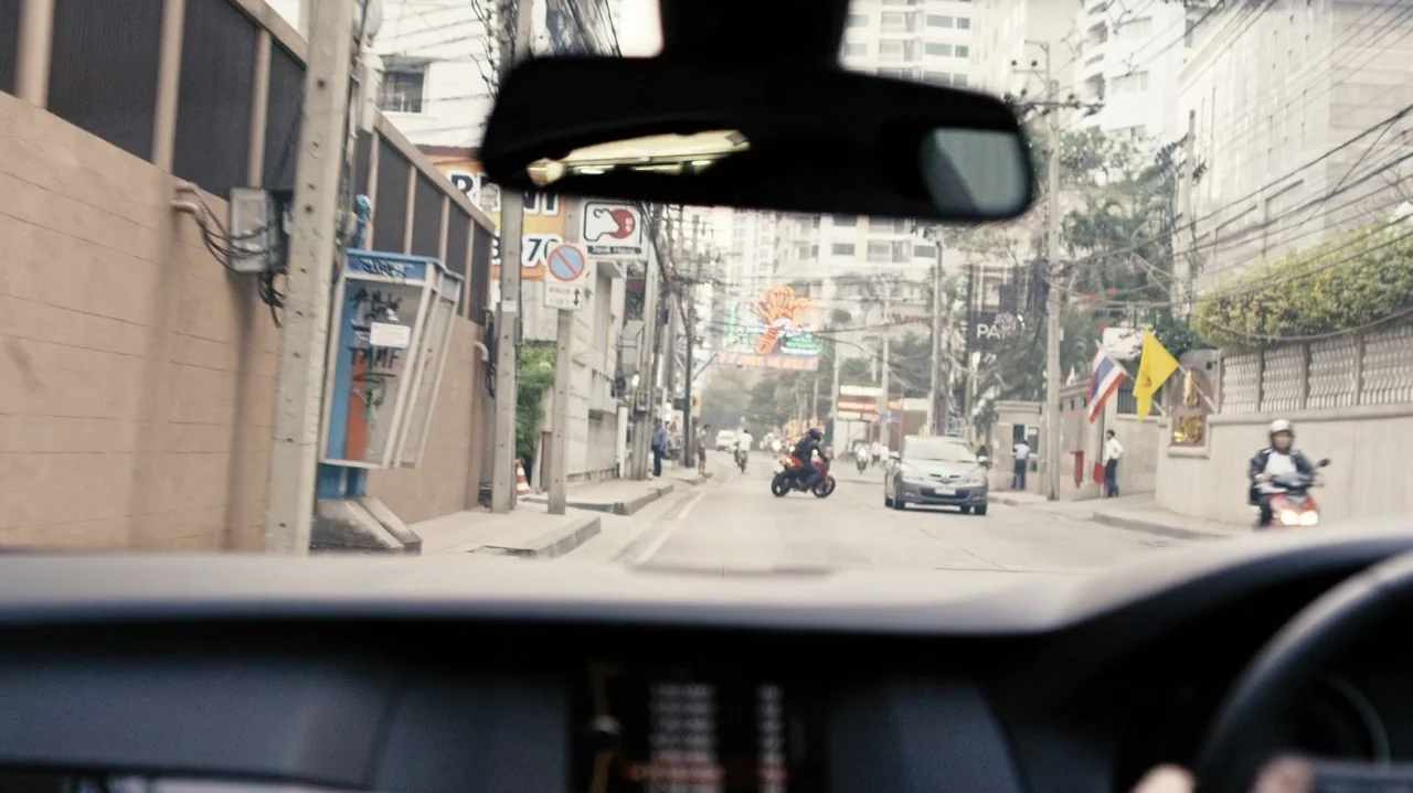 Zero Tolerance (2015) movie still: wide — A dense urban street in Thailand lined with concrete walls and utility poles. Multiple vehicles and…