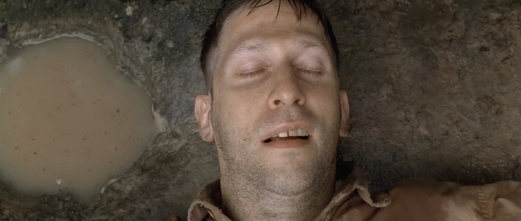 O Brother, Where Art Thou? (2000) movie still: closeup — A man lies on the damp, textured ground beside a small pool of muddy water. His face is…