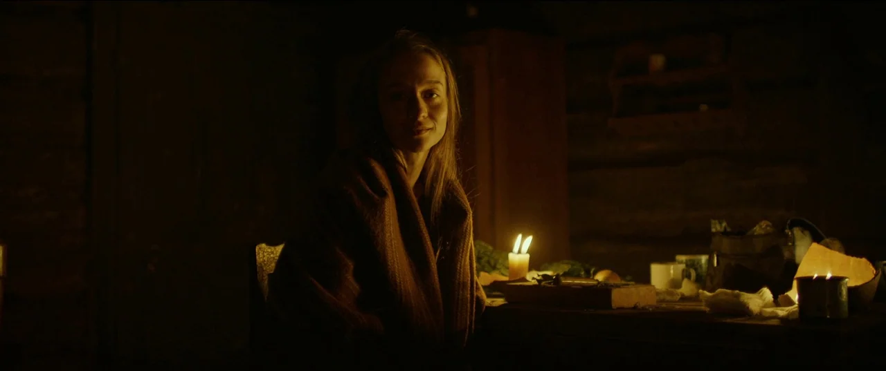 Nightsiren (2022) movie still: medium — A dark wooden interior is lit by a flickering candle on a rough table. A woman sits at the table wearing a…