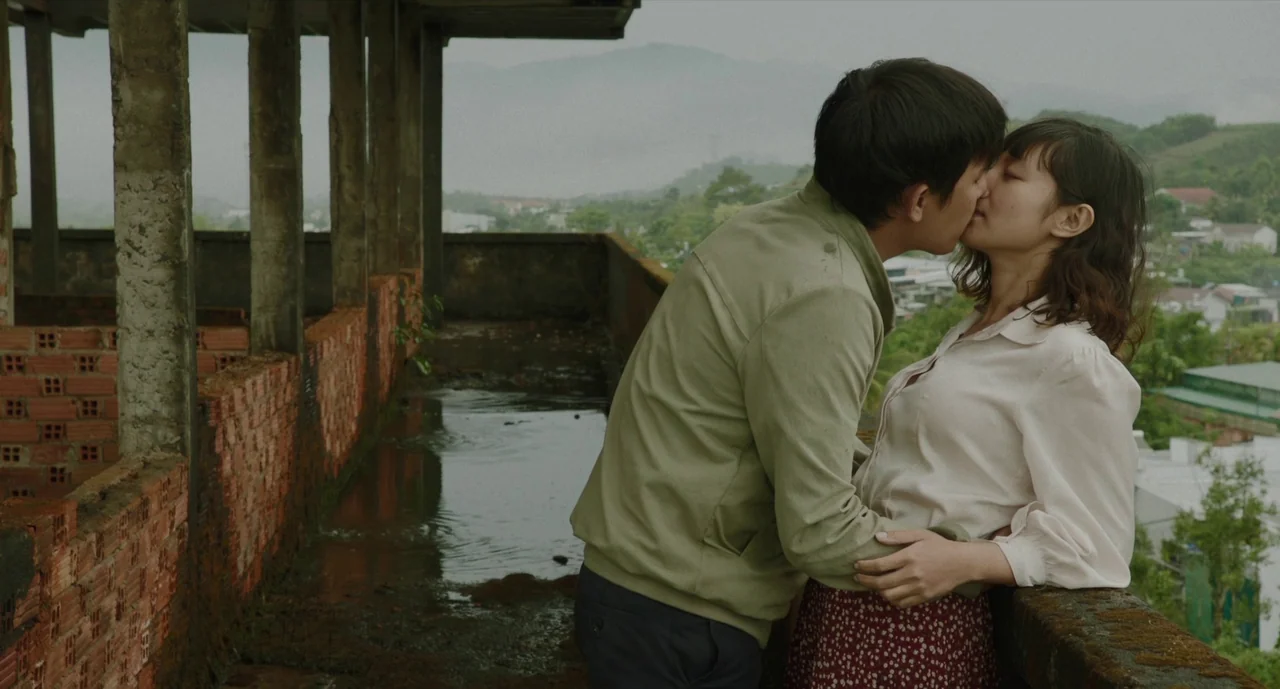 Inside The Yellow Cocoon Shell (2023) movie still: medium — A partially constructed concrete building overlooks a misty, green rural valley. A young…