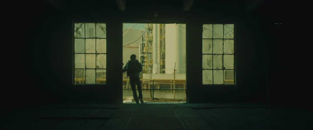 A Most Violent Year (2014) movie still: wide — A dark, empty industrial space with large paned windows faces an outdoor refinery area. A solitary…