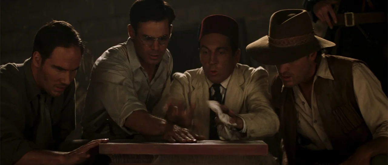 The Mummy (1999) movie still: medium — A dimly lit, underground stone chamber features dark masonry and an inscribed slab. Four men in 1930s attire…