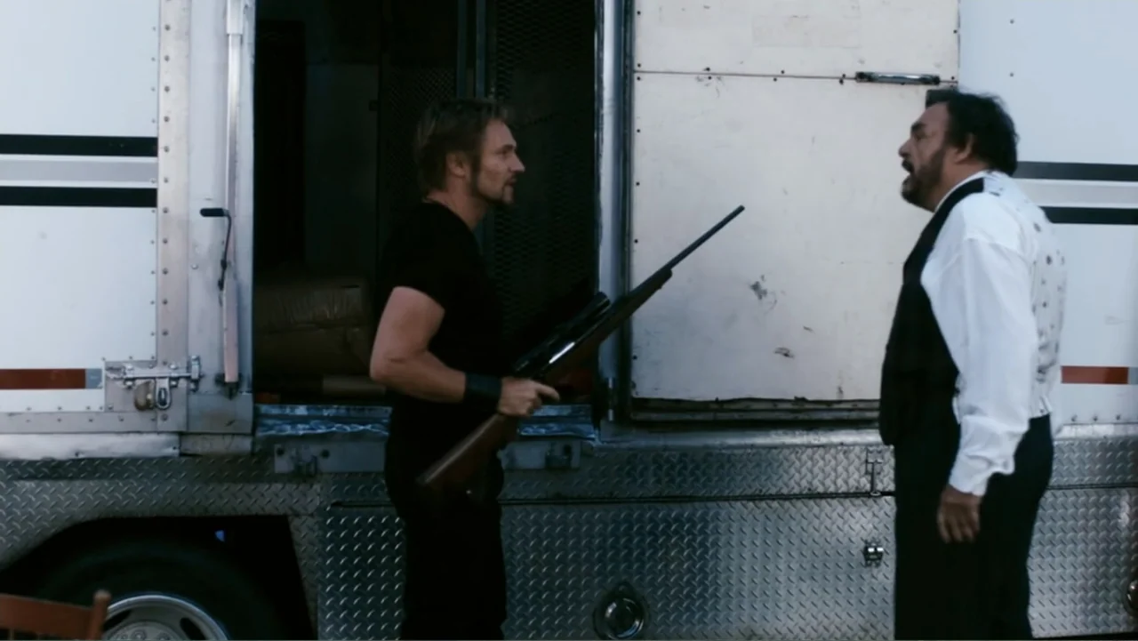 Sophie & Sheba (2010) movie still: fullbody — An outdoor area next to a white metal trailer with a checkered pattern. Two men converse while one…