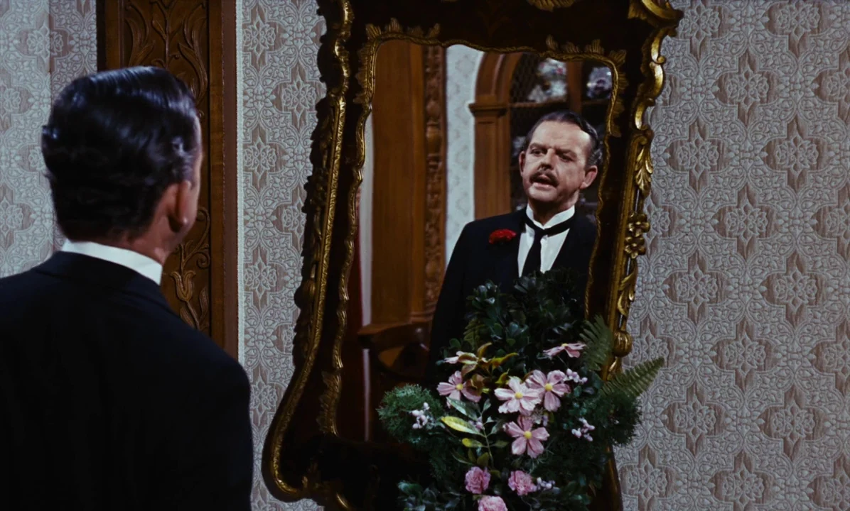 Mary Poppins (1964) movie still: medium — An ornate mirror hangs on a wall covered in patterned 1950s beige wallpaper. A man in a suit looks at his…