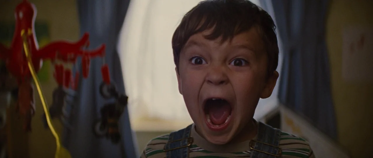 Looper (2012) movie still: closeup — A dimly lit room with a hanging mobile visible in the foreground. A young boy stands facing the camera with his…