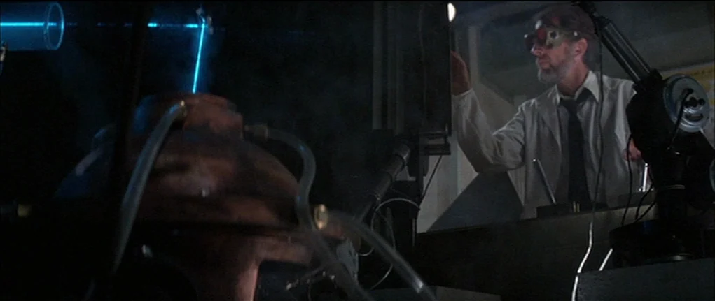 Demon Seed (1977) movie still: medium — A dimly lit scientific workspace features a metal apparatus in the immediate foreground. An older man in a…
