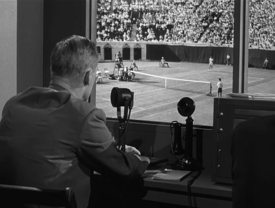 Strangers on a Train (1951) movie still: medium — A man in a suit sits in a broadcast booth overlooking a tennis match. A large window provides a…