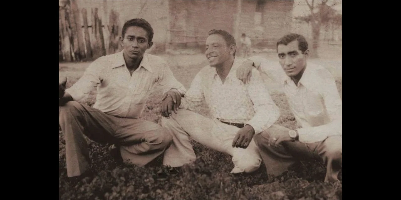 Leyenda Viva (2022) movie still: medium — A group of three men are kneeling in a dirt field. The men wear light-colored collared shirts and trousers…