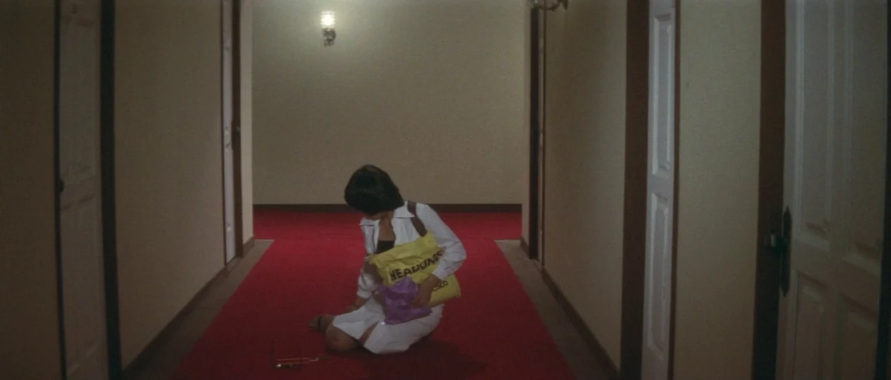 Take Me Away! (1978) movie still: fullbody — A long, symmetrical indoor corridor with beige walls and a vibrant red runner carpet. A person kneels…