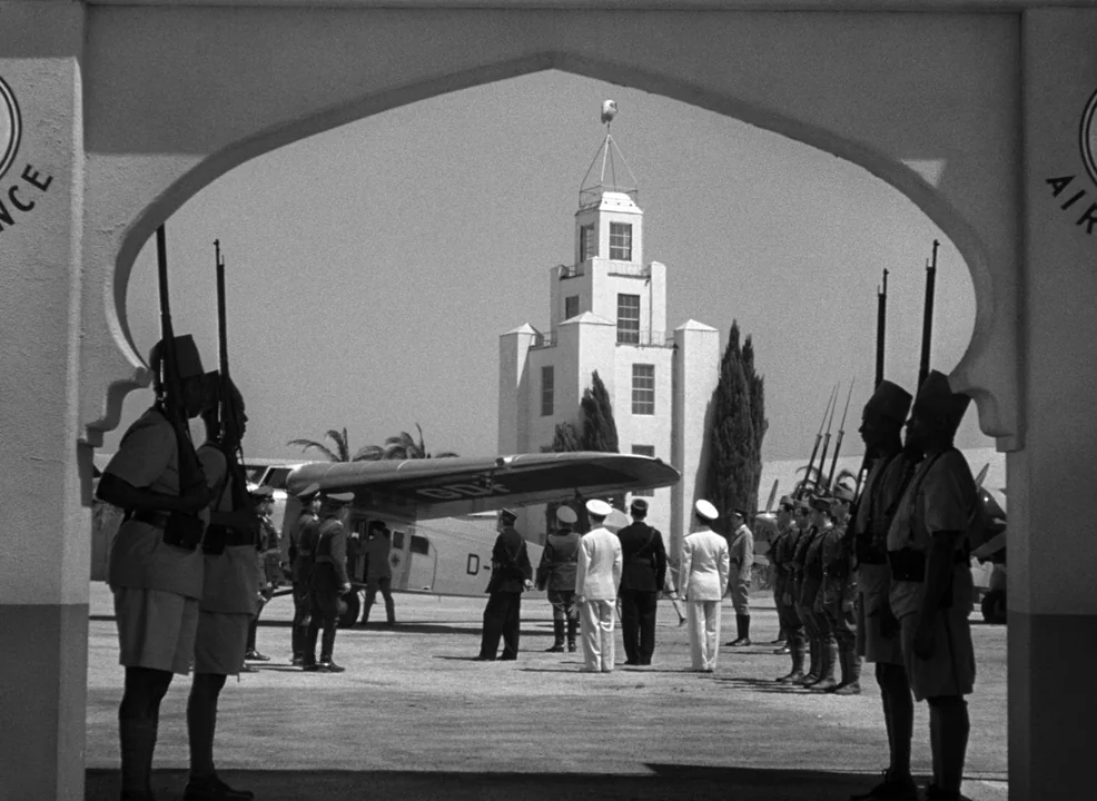 Casablanca (1942) movie still: wide — An airfield scene framed through a Moorish-style archway features a vintage airplane. Soldiers stand in…