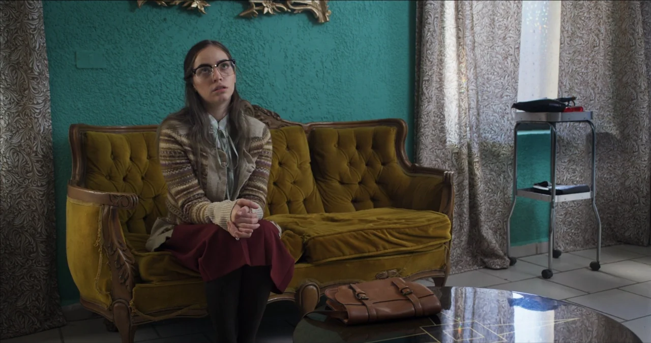 Bendita Suegra (2023) movie still: medium — A room with textured teal walls and a vintage mustard-yellow velvet tufted sofa. A young woman with…
