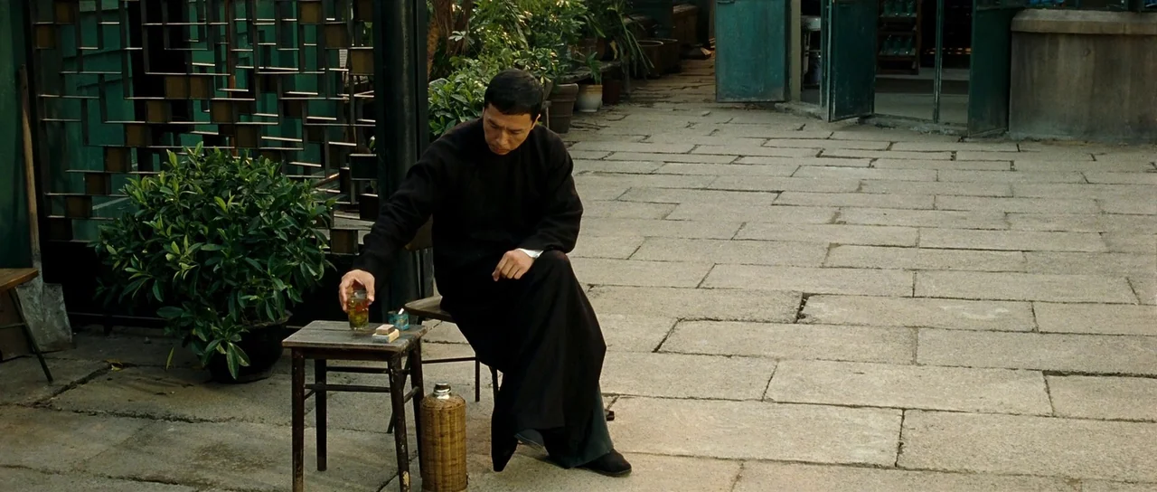 Ip Man 2 (2010) movie still: fullbody — A stone-paved courtyard features potted greenery and a traditional metal-barred divider. A man in a dark…