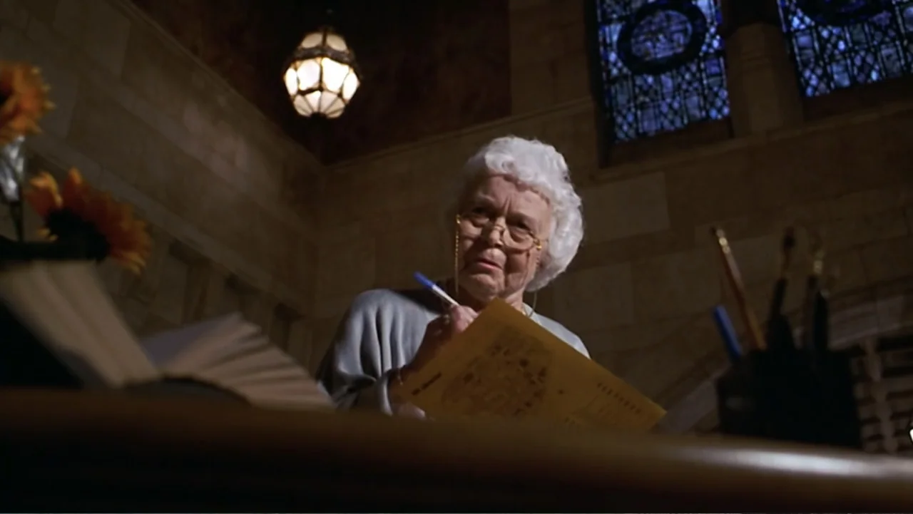Matilda (1996) movie still: medium — An interior space with arched stone architecture and a stained glass window. An elderly woman sits at a desk,…