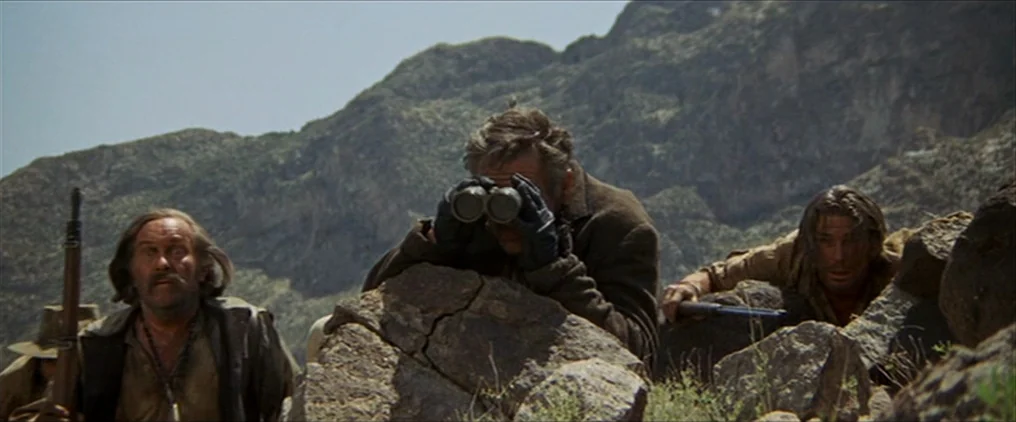 The Wild Bunch (1969) movie still: medium — Three men crouch among large boulders and sparse vegetation in a mountainous landscape. The men hold…