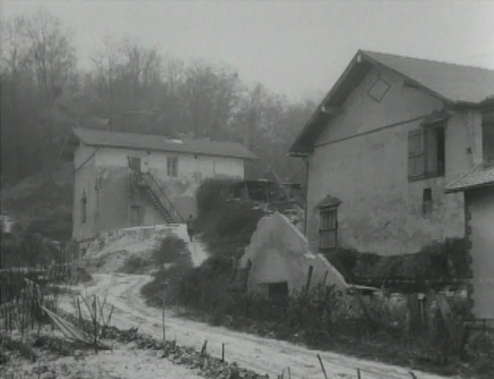 Les Carabiniers (1963) movie still: establishing — A rural setting features aging stone houses situated on a muddy, unpaved path. Bare trees frame…