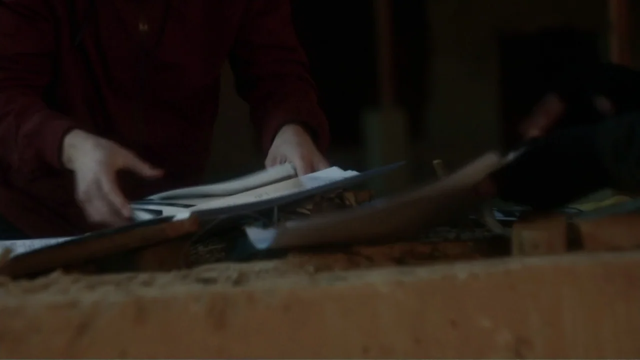 Code of Trust (2019) movie still: closeup — A workbench cluttered with wooden shavings and documents rests in a dimly lit room. Hands reaching into…
