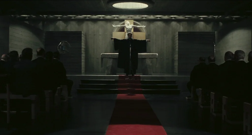 Todo Modo (1976) movie still: wide — A dark concrete chapel features a central red carpet aisle and a wall sculpture. A man in black religious…