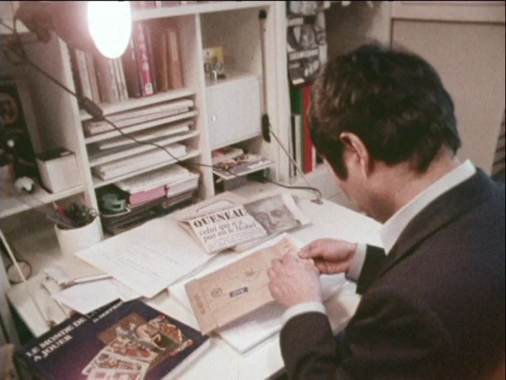 Catalogue of Memory (1977) movie still: medium — A cluttered white desk holds several books, a lamp, and various stacks of paper. A man in a dark…