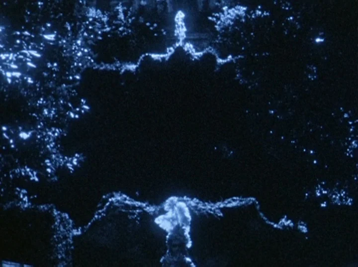 Eaux d’Artifice (1953) movie still: establishing — A dark garden space is illuminated by high-contrast blue light at night. A central fountain…