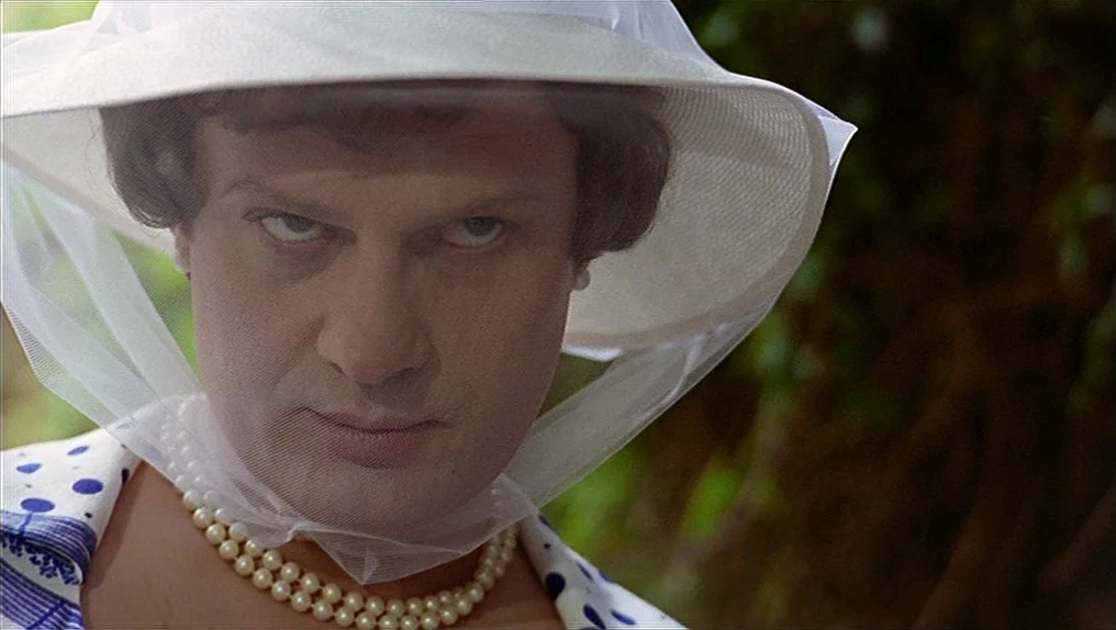 In A Year Of Thirteen Moons (1978) movie still: closeup — A person stands outdoors against blurred green leaves wearing a large brimmed hat. The…