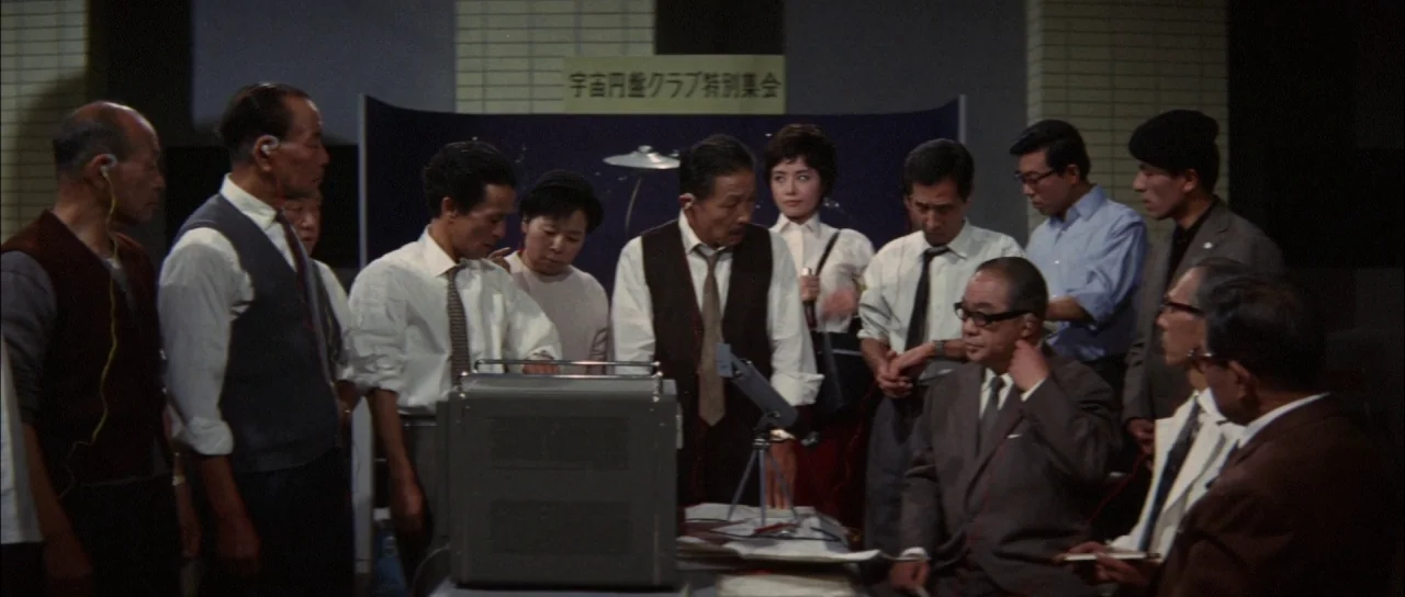 Ghidorah The Three Headed Monster (1964) movie still: medium — A group of people are standing and sitting around a table in a room. They examine a…