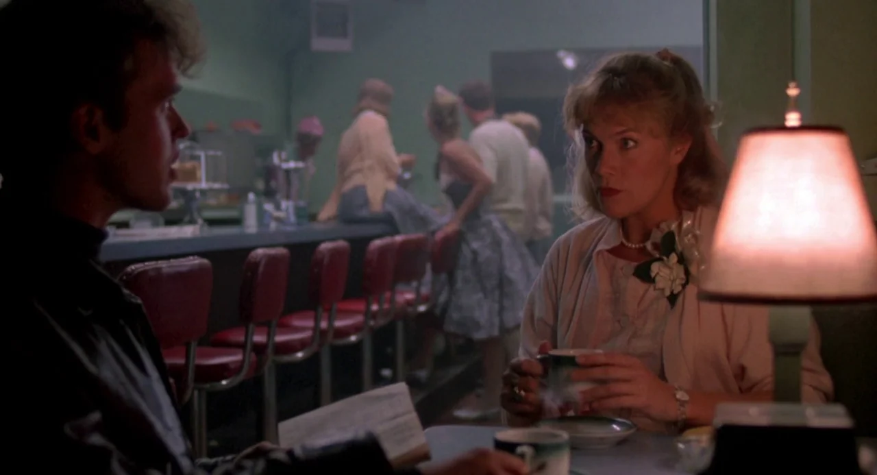 Peggy Sue Got Married (1986) movie still: medium — A 1950s diner booth with red vinyl stools and a green-walled background. A man and woman hold…