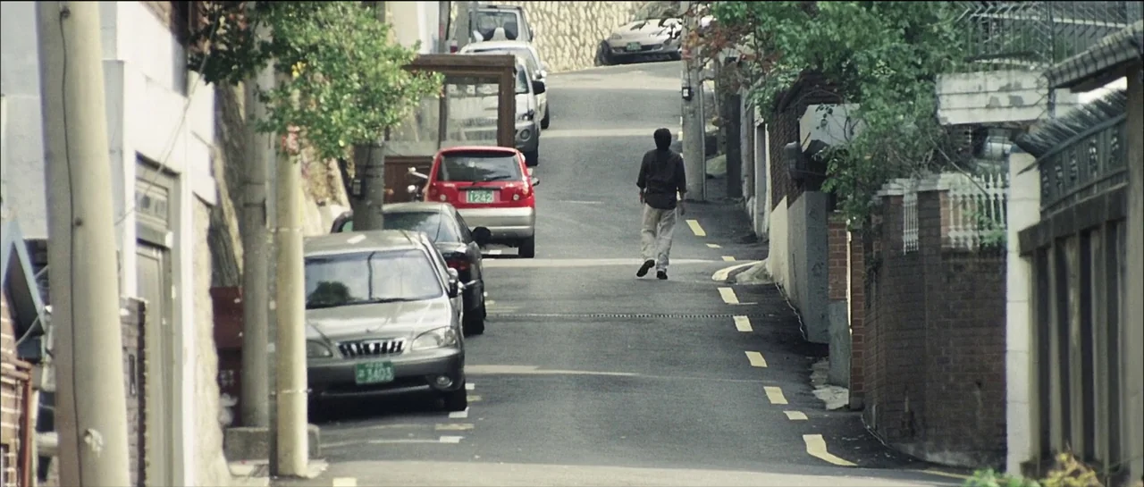 The Chaser (2008) movie still: wide — A narrow residential road slopes uphill between brick walls and parked cars. A lone man walks away from the…