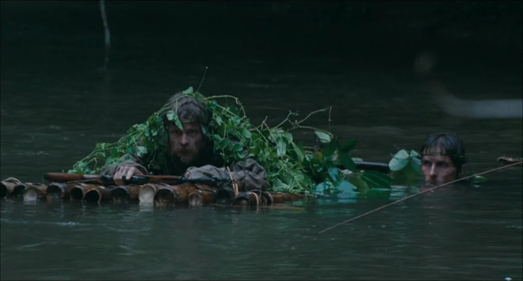 Rescue Dawn movie still: medium — A murky, slow-moving river flows through a dense and dark environment with thick vegetation. Two men in tactical…