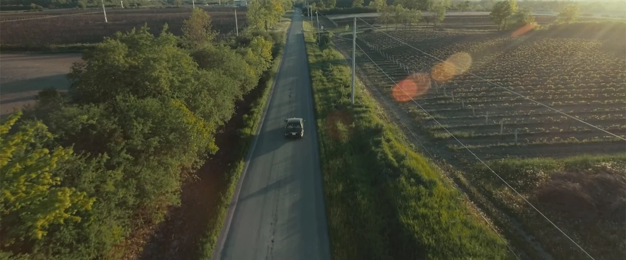 Jump, Darling (2022) movie still: wide — A long asphalt road stretches through flat rural farmland bordered by green trees. A dark sedan drives down…