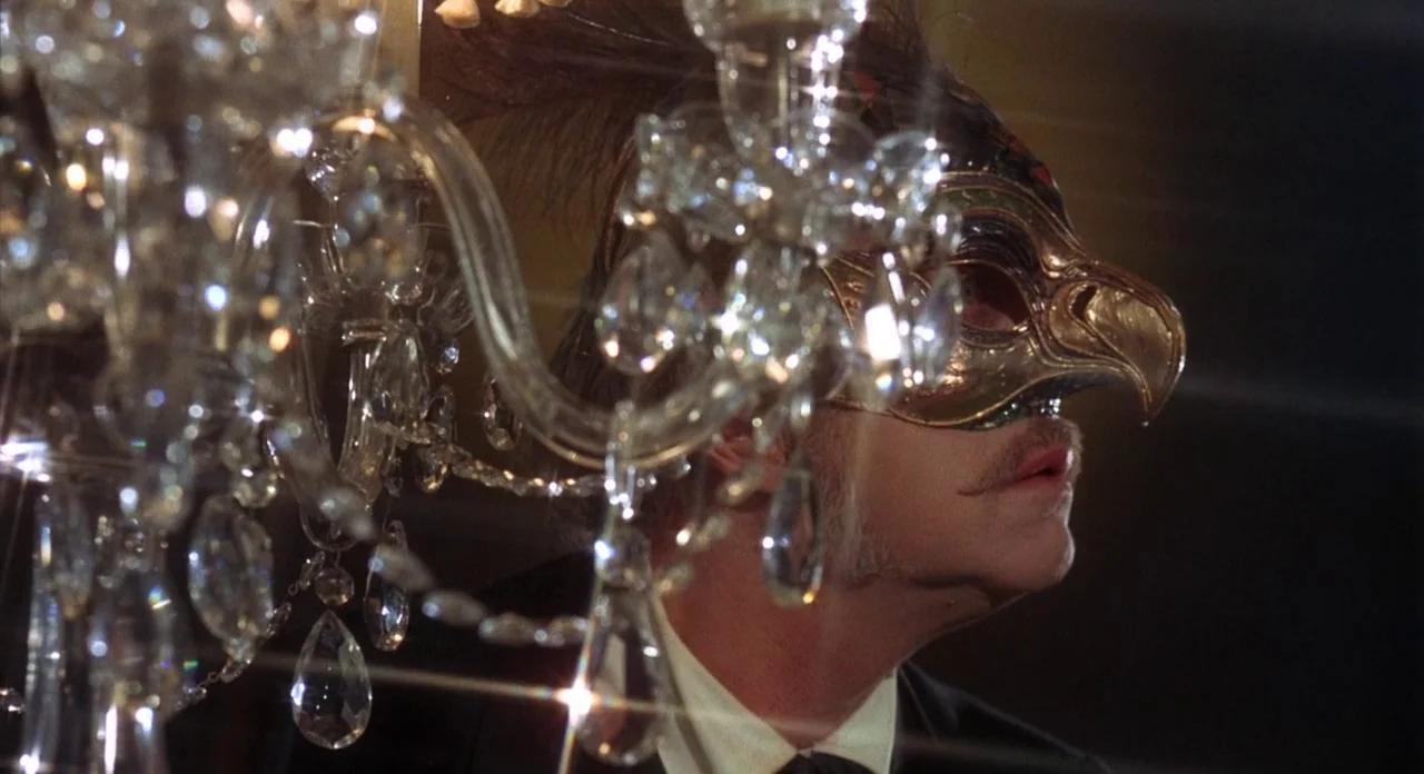 The Abominible Dr. Phibes (1972) movie still: closeup — A man wears an ornate gold bird mask during an evening event. Crystal droplets from a large…