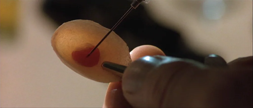 Gattaca (1997) movie still: closeup — A close-up view shows a needle piercing a small, pale, translucent, oval object. Fingers hold the object…