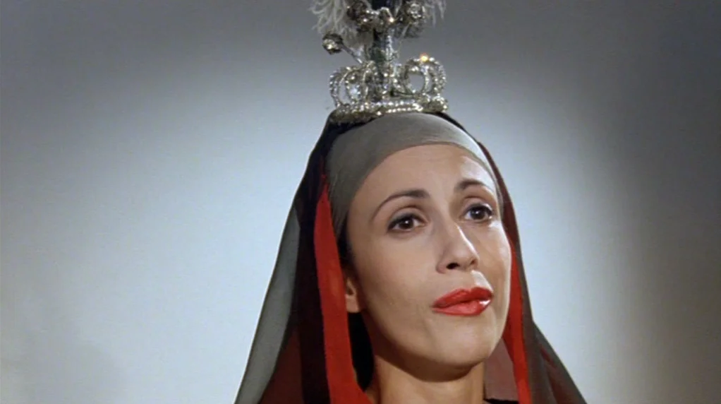 Malina (1991) movie still: closeup — A woman wears an elaborate, jeweled crown and head covering against a plain background. She stands facing…
