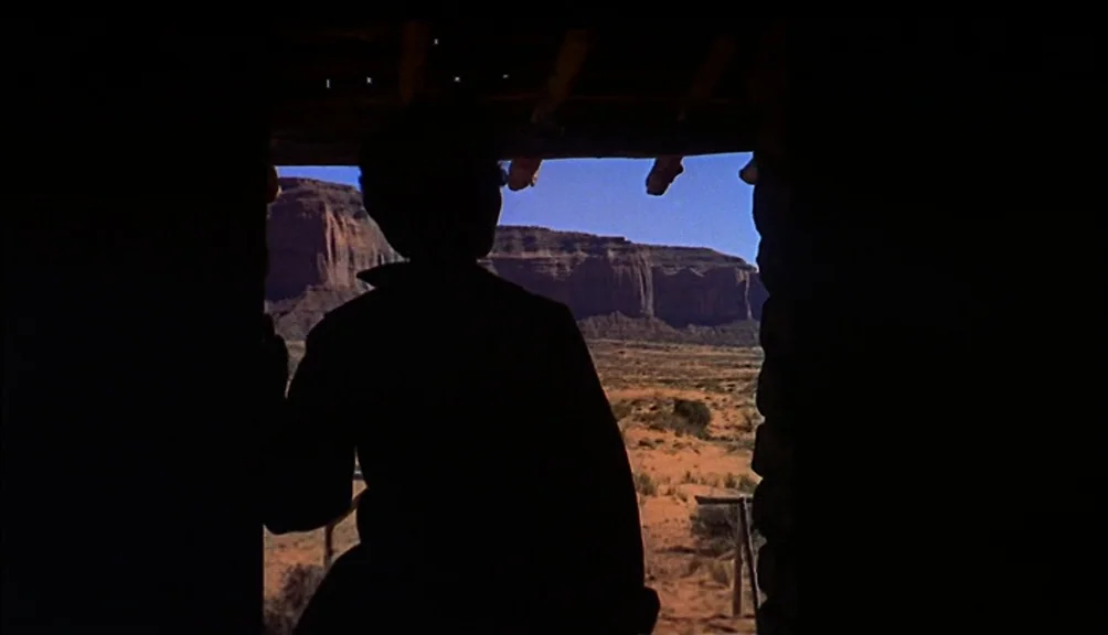 The Searchers movie still: medium — A dark wooden doorway frames a brightly lit arid desert with large sandstone mesas. A silhouette of a person…