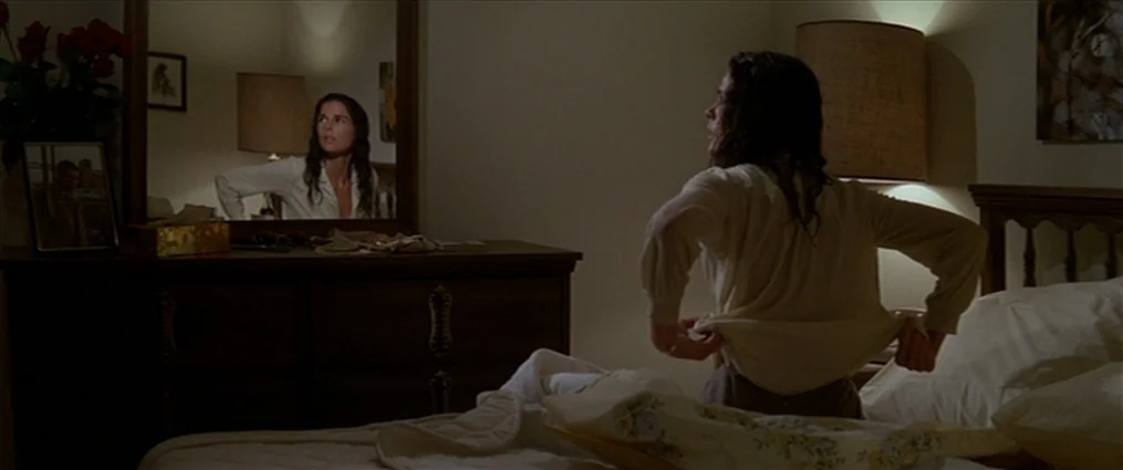 The Getaway (1972) movie still: medium — A dimly lit room contains a bed and a wooden vanity with a mirror. A woman wearing a cream shirt sits on…