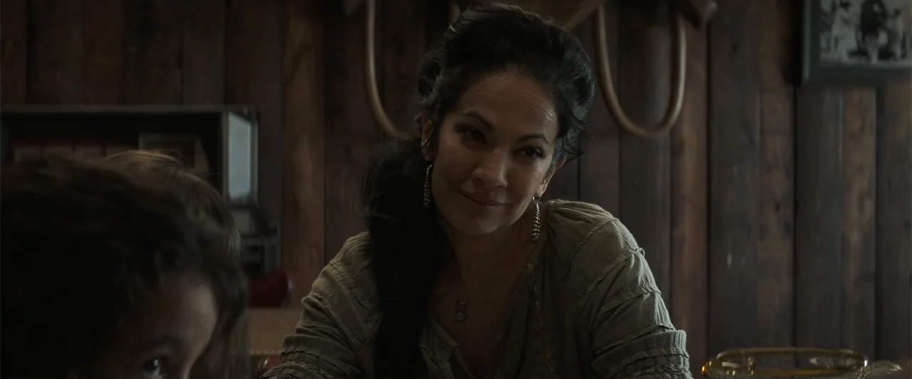 Cry Macho (2021) movie still: medium — A rustic interior features dark vertical wood plank walls and minimal decor. A woman with dark hair sits…