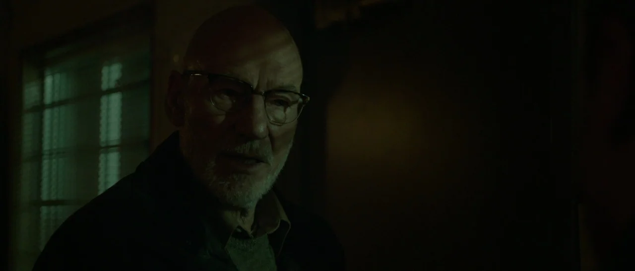 Green Room (2015) movie still: closeup — An interior space is dimly lit by light filtering through slatted blinds. A bald, elderly man stands in the…