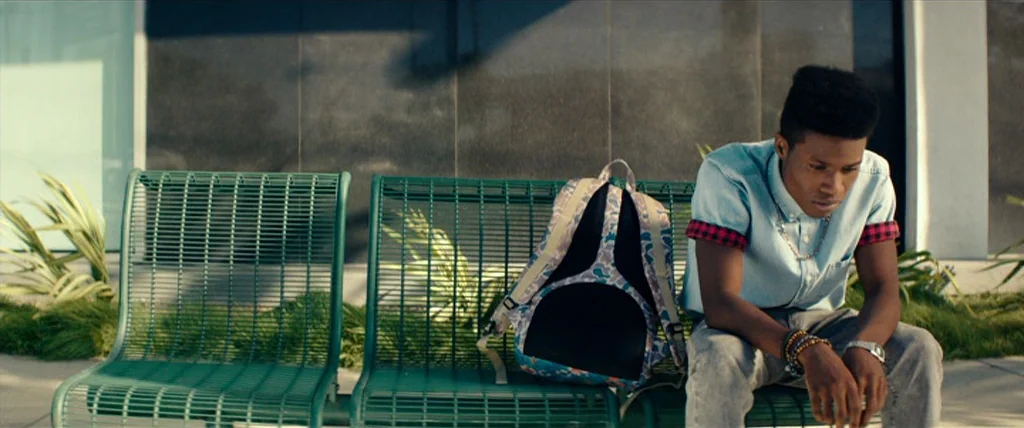 Dope (2015) movie still: medium — A green metal bench sits against a dark stone wall in an outdoor area. A young man sits on the right side of the…