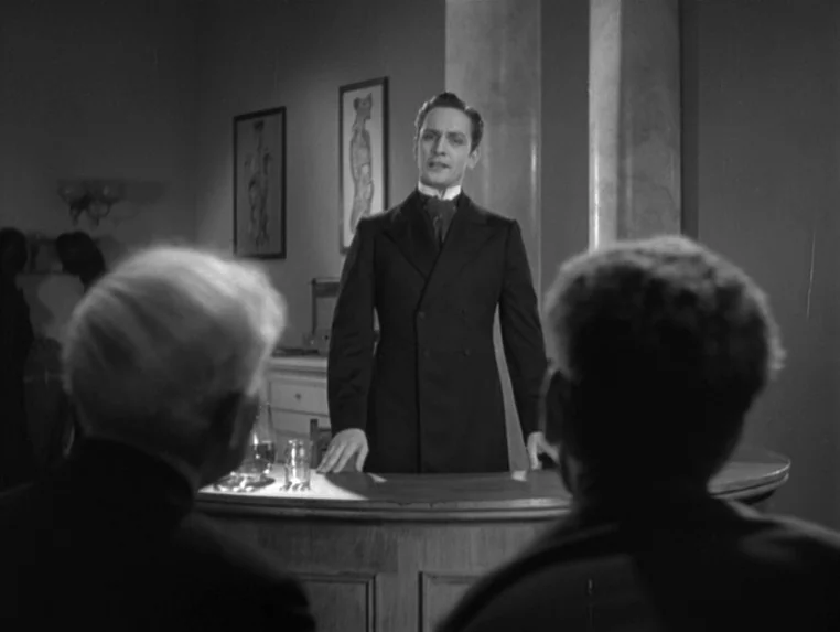 Dr. Jekyll and Mr. Hyde (1931) movie still: medium — A wood-paneled room features medical anatomical diagrams on the wall behind a desk. A man in a…