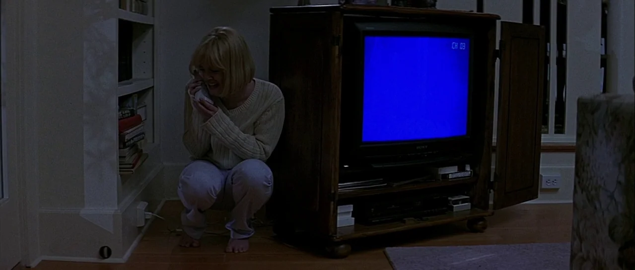 Scream (1996) movie still: medium — A dimly lit room features a woman crouched against a wooden cabinet containing a television. The blue light from…