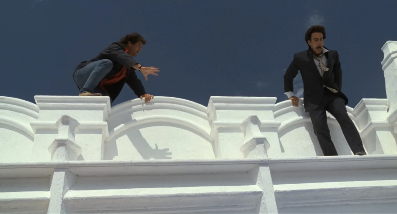 Lethal Weapon (1987) movie still: wide — Two men are balanced on a stark white curved roof ledge against a blue sky. One man reaches out toward the…