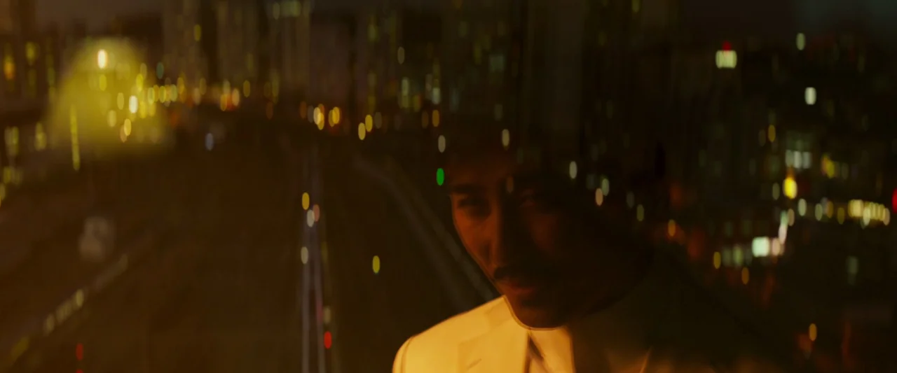 Believer (2018) movie still: closeup — A man is positioned against a large glass window overlooking a blurred nighttime city. The view through the…