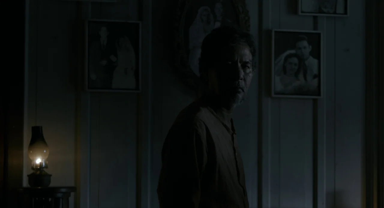 In My Mother’s Skin (2023) movie still: medium — A dimly lit room with vertical wood paneling and framed wedding portraits on walls. An elderly man…