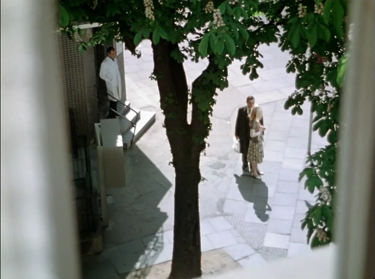 Fear Of Fear (1975) movie still: wide — A paved area surrounds a large central tree with green leaves and blossoms. Three people stand at different…