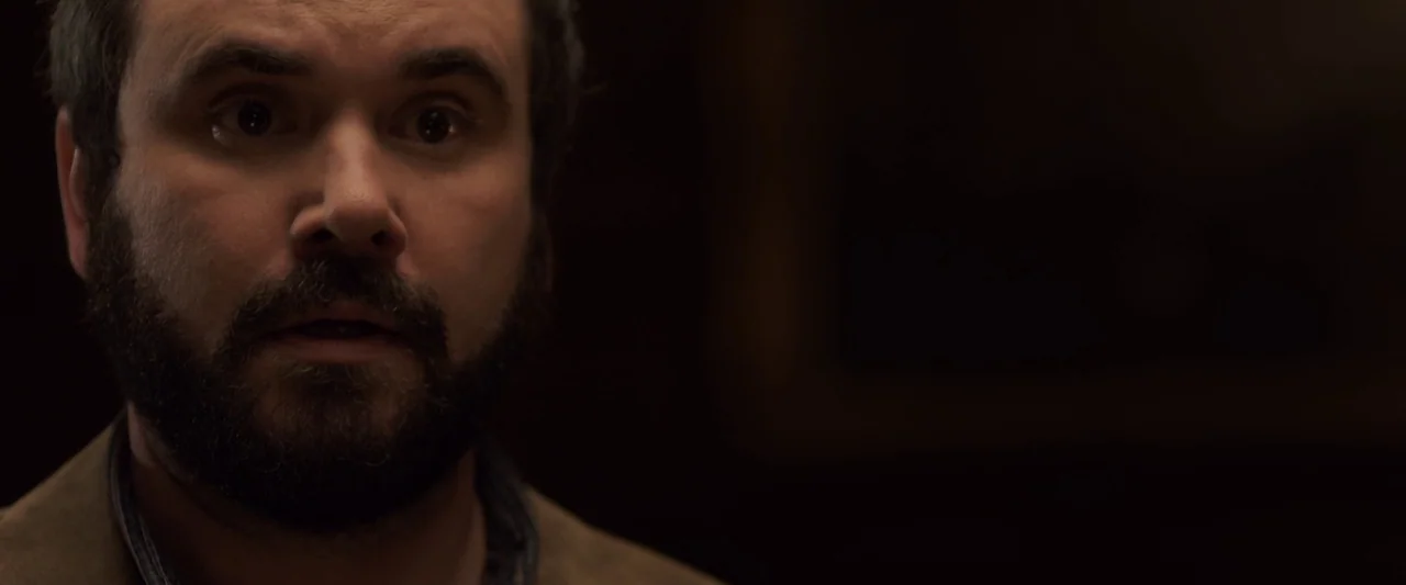 You’re Next (2013) movie still: closeup — A man stands in a poorly lit, dark interior space with shadowed surroundings. He stares forward with an…