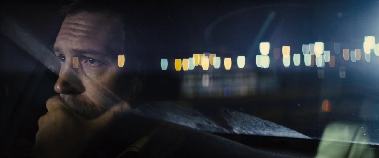Locke (2013) movie still: closeup — A man sits inside a vehicle at night with blurred city lights visible through glass. The man looks out the side…